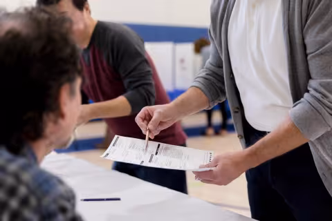 Person standing and reviewing a ballot in the background as a person sits in the foreground.