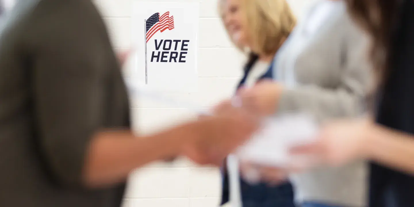"Vote here" sign in focus in the background with people in the foreground out of focus exchanging papers 